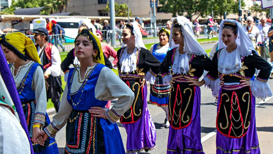 Young dancers in embroidered folk dress, white veils and purple skirts wait in the sun before a street parade