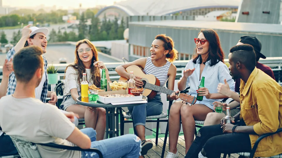 Friends of many backgrounds laugh around outdoor tables while one plays a guitar