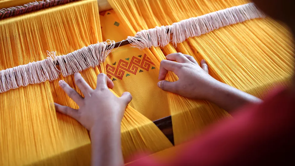 Hands weave a pattern into bright yellow threads on a wooden loom