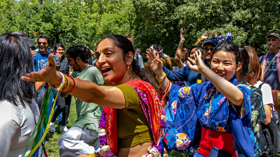 A woman in a sari and a woman in a blue yukata dance and laugh in a sunny park crowd