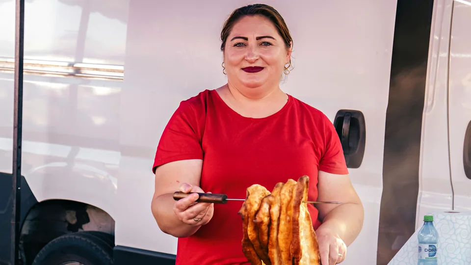 A smiling cook in a red T-shirt holds up a skewer of golden fried dough at her food stall