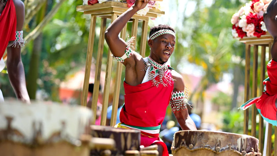 A drummer in red with beaded bands dances between big hand drums at an outdoor festival