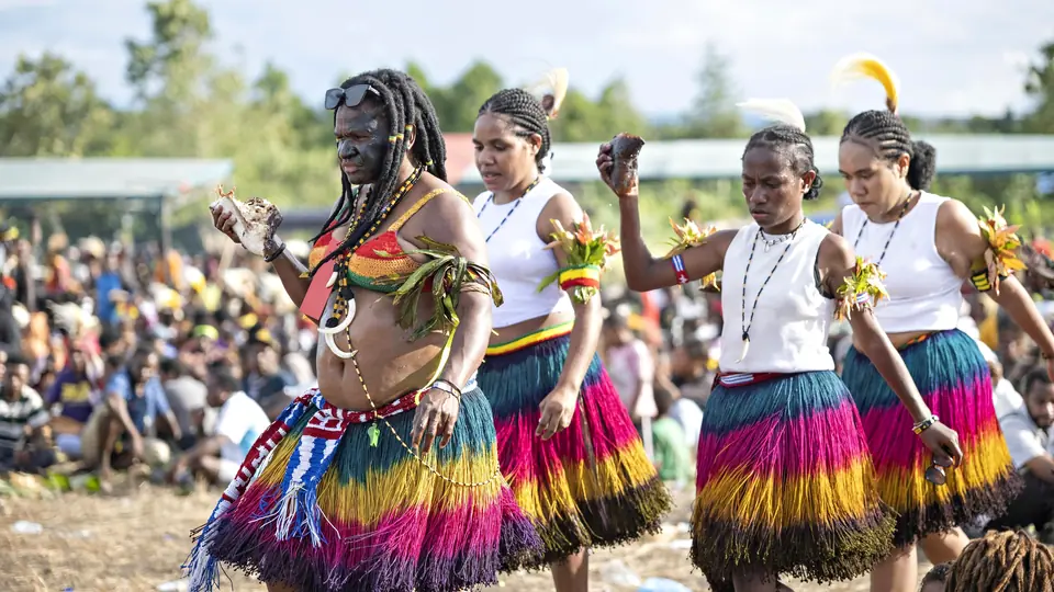 Dancers in striped grass skirts and feathers perform for a large outdoor crowd