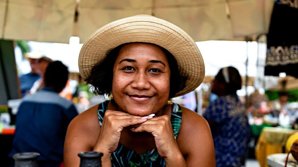 A smiling woman in a straw hat rests her chin on her hands at a market stall