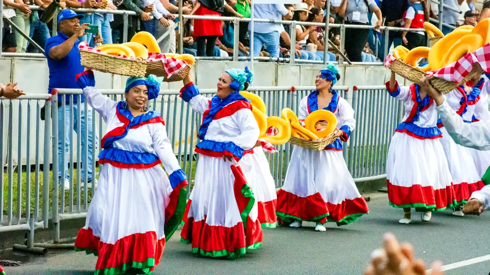 Dancers in white, red and blue skirts parade with baskets of bread past a full stand of spectators