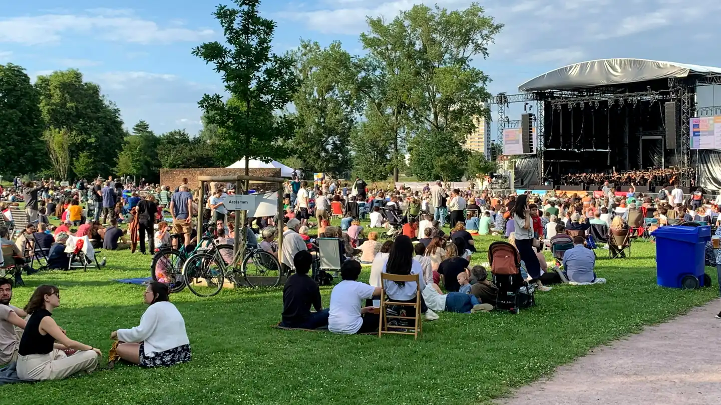 Hundreds of people fill a green park lawn in front of an open-air stage on a sunny day