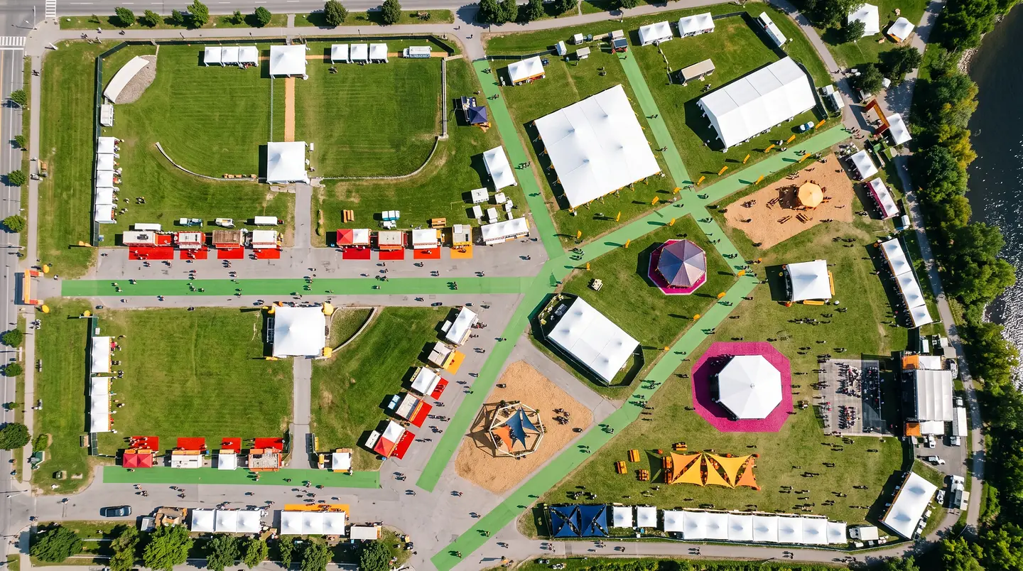 A straight-down aerial of a festival ground: white marquees, rows of food trucks, green walkways, a sand play area and two coloured circus tents laid out between the roads and the river.